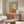 May include: Framed poster artwork displayed above wooden sideboard in modern living room. The painting shows a white fluffy terrier dog wearing yellow goggles and a flowing pink-coral scarf against sage green background. Mid-century furniture includes walnut credenza, white curved chair, oval glass coffee table, and brass floor lamp in neutral-toned space with natural lighting.