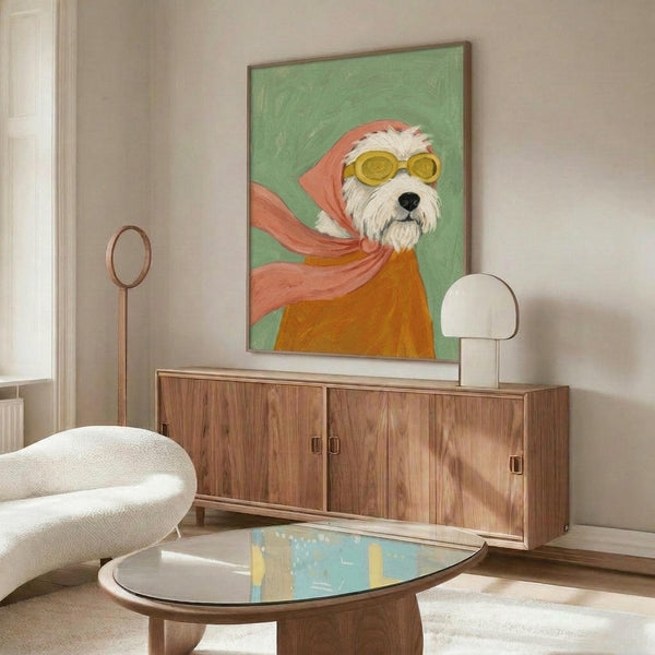May include: Framed poster artwork displayed above wooden sideboard in modern living room. The painting shows a white fluffy terrier dog wearing yellow goggles and a flowing pink-coral scarf against sage green background. Mid-century furniture includes walnut credenza, white curved chair, oval glass coffee table, and brass floor lamp in neutral-toned space with natural lighting.