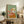 May include: Framed artwork displaying a white terrier dog wearing yellow goggles and a flowing pink scarf against a sage green background, styled in painterly brushstrokes with warm orange and coral tones. The piece sits on a rustic wooden console alongside books, pottery, and a fern plant in a cozy living room setting with cream walls and brown leather furniture.