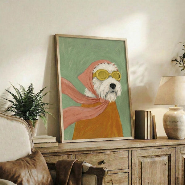 May include: Framed artwork displaying a white terrier dog wearing yellow goggles and a flowing pink scarf against a sage green background, styled in painterly brushstrokes with warm orange and coral tones. The piece sits on a rustic wooden console alongside books, pottery, and a fern plant in a cozy living room setting with cream walls and brown leather furniture.
