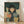 May include: Framed art print leaning against white wall on light wood floor. Illustration shows young girl with black bob haircut and rosy cheeks wearing navy polka dot dress, surrounded by eight cats in various colors - orange tabby, gray, white, blue-gray, black, and cream. Dark teal background with vertical rain lines and scattered leaves creates moody atmosphere. Natural wood frame visible.