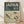 May include: Framed vintage-style travel poster with dark green header and cream background, featuring four colorful birds perched on cherry blossom branches with pink flowers, displayed against white textured wall on light wood floor with natural lighting creating soft shadows