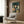 May include: Framed poster artwork displayed on wood-paneled wall above beige upholstered armchair with geometric pillow. Artwork shows young girl with black bob haircut surrounded by multiple cats in various colors including orange, gray, white and black, set against teal background with falling rain. Modern reading nook with brass floor lamp and side table.
