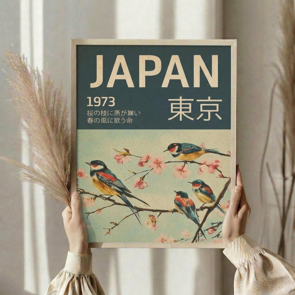 May include: Hands holding a framed vintage-style travel poster featuring four colorful birds perched on cherry blossom branches against a soft green background. The poster displays "JAPAN 1973" and Japanese characters in cream lettering at the top. The birds have blue, orange, and yellow plumage. A bundle of dried pampas grass is visible on the left side, and white curtains create a soft backdrop in this bright, naturally-lit room setting.