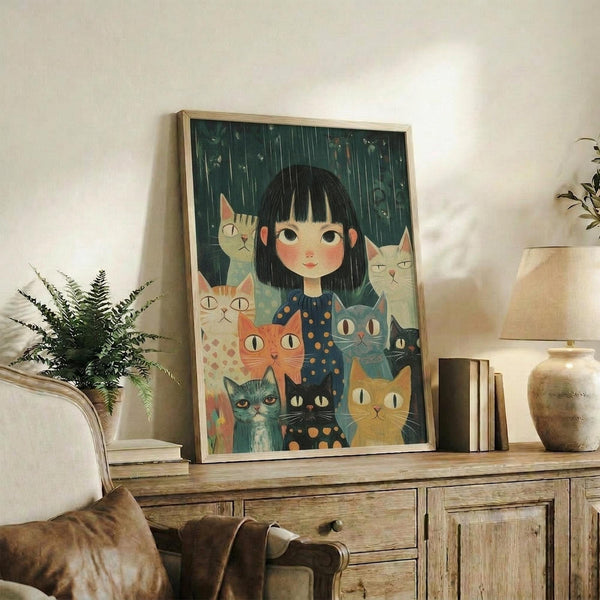 May include: Framed artwork displaying a young girl with black bob haircut surrounded by seven colorful cats in various shades of orange, gray, blue, and black against a dark green rainy background. The illustration has a whimsical, folk art style with the girl wearing a dark blue polka dot dress. The wooden-framed print sits on a rustic wooden sideboard alongside books, a ceramic vase, and a fern plant, with a beige armchair visible in the cozy living room setting.