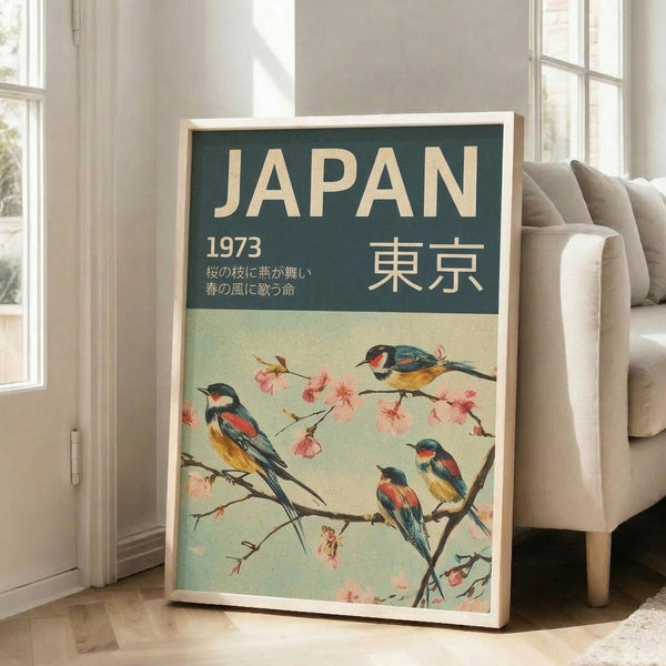 May include: A vintage-style travel poster in a light wood frame leaning against a white wall beside a beige sofa. The poster features a dark green header with cream text reading "JAPAN 1973" and Japanese characters, below Japanese text. The main image shows four colorful birds with red, blue, and yellow plumage perched on cherry blossom branches with delicate pink flowers against a soft mint green background.