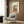 May include: Framed poster print of a charming two-story Japanese building with blue-green tiled roof, warm beige walls, round window, small balcony, and bicycle parked outside. Power lines and utility pole visible. Artwork displayed above a beige upholstered armchair with geometric patterned pillow in a modern room with wood paneling, floor lamp, and side table with open book.