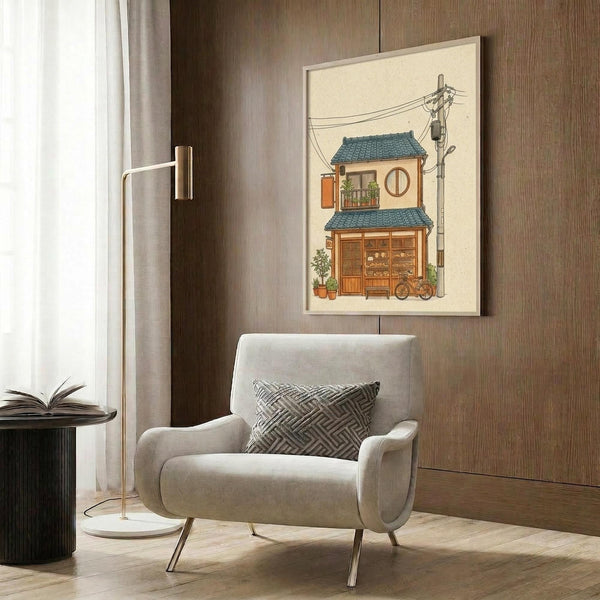 May include: Framed poster print of a charming two-story Japanese building with blue-green tiled roof, warm beige walls, round window, small balcony, and bicycle parked outside. Power lines and utility pole visible. Artwork displayed above a beige upholstered armchair with geometric patterned pillow in a modern room with wood paneling, floor lamp, and side table with open book.