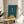 May include: Minimalist artwork in wooden frame showing geometric bicycle handlebars and front wheel in teal blue and mustard yellow colors, displayed on round wooden side table next to beige upholstered chair with white throw pillow, green plant leaves visible, soft natural lighting creating shadows on white wall