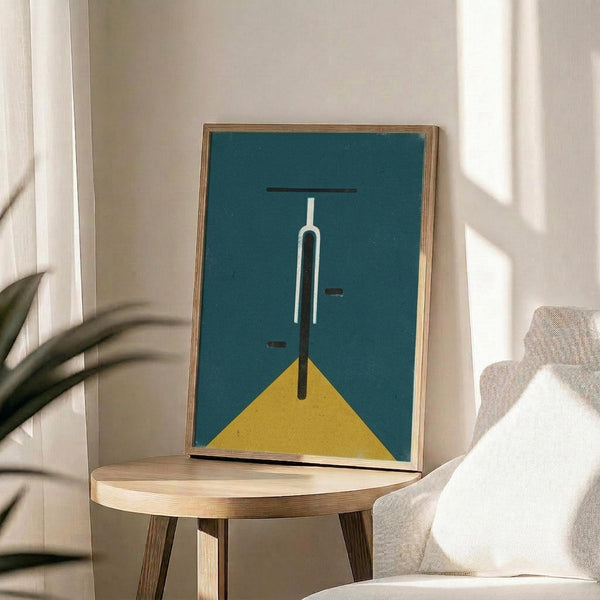 May include: Minimalist artwork in wooden frame showing geometric bicycle handlebars and front wheel in teal blue and mustard yellow colors, displayed on round wooden side table next to beige upholstered chair with white throw pillow, green plant leaves visible, soft natural lighting creating shadows on white wall