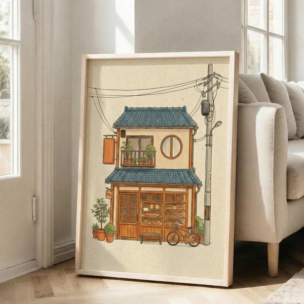 May include: Framed illustration of a charming two-story Japanese bakery with teal tile roof, warm wooden storefront displaying bread and pastries, small balcony with plants, bicycle parked outside, and overhead power lines. The artwork has a cream background and is displayed in a light wood frame leaning against a white wall next to a beige sofa in a bright, modern living room with large windows.