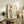 May include: Framed art print displayed on rustic wooden console table showing Japanese woman in flowing green kimono with coral obi sash, holding red katana sword. Soft watercolor style with muted background. Scene includes fern plant, ceramic vase, stacked books, and cream upholstered chair in warm-toned living room setting.