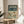 May include: Framed vintage-style travel poster displayed on round wooden side table next to beige upholstered chair. Poster features teal header with cream text reading "JAPAN 1973" and Japanese characters, below colorful illustrated birds perched on cherry blossom branches against pale green background. Natural lighting creates soft shadows on white wall behind wooden frame.