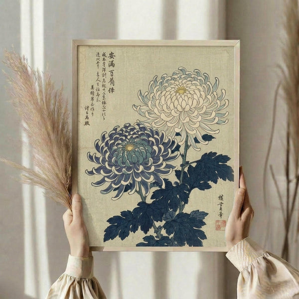May include: Hands holding a framed botanical print featuring two chrysanthemum flowers - one dark blue-purple and one light cream colored - with dark blue-green leaves and Japanese calligraphy text. The artwork has a vintage beige background and is displayed in a light wood frame. Dried pampas grass stems are held alongside the frame against soft neutral curtains and natural window lighting.