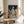 May include: Framed art print displayed on round wooden side table next to beige upholstered chair. Artwork shows woman with red hair in profile wearing blue and white leaf-patterned dress, surrounded by orange and peach magnolia flowers against dark teal background. Natural wood frame, soft window lighting, plant leaves visible in corner.