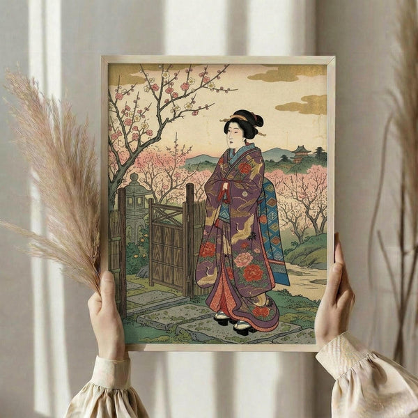 May include: Hands holding a framed Japanese woodblock print featuring a woman in an ornate purple kimono with floral patterns standing near cherry blossom trees. The artwork shows traditional ukiyo-e style with muted colors and detailed landscape background including mountains and buildings. A bundle of dried pampas grass sits nearby against white curtains in soft natural lighting.