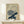 May include: Vintage Japanese botanical print in light wood frame showing two detailed chrysanthemum flowers - one purple-blue and one cream colored - with dark blue-green leaves against beige background, displayed on light wood console table with small white wrapped gift beside it on white wall.