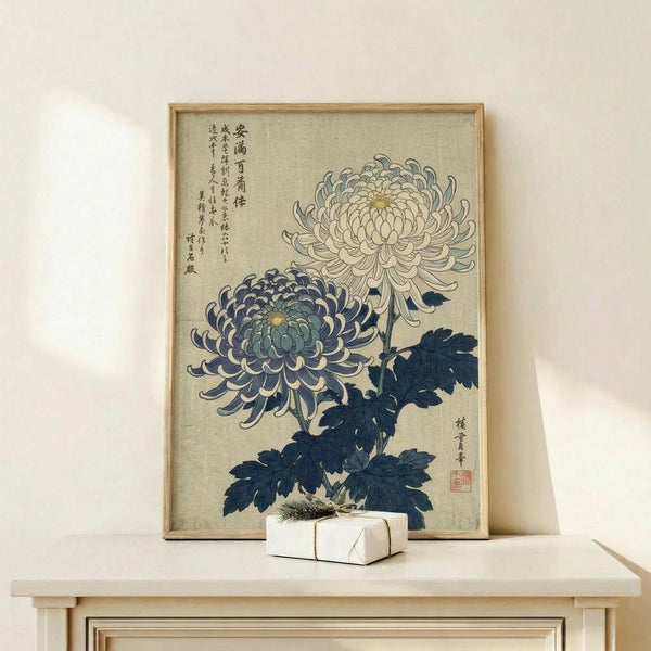 May include: Vintage Japanese botanical print in light wood frame showing two detailed chrysanthemum flowers - one purple-blue and one cream colored - with dark blue-green leaves against beige background, displayed on light wood console table with small white wrapped gift beside it on white wall.