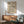 May include: Mid-century modern living room with sage green armchair and wood credenza. Framed geometric poster with cream background and black diamond pattern, displayed above sideboard. Table lamp with white shade and small round coffee table visible. Natural sunlight creates shadows on white wall.