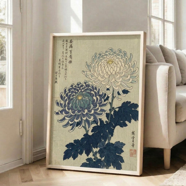 May include: Framed Japanese botanical print featuring two large chrysanthemums - one white and one blue-purple - with dark blue leaves on beige background. Asian calligraphy text visible in upper left corner with red artist seal. Light wood frame leaning against beige chair in bright, minimalist living room with white walls and natural lighting from windows.