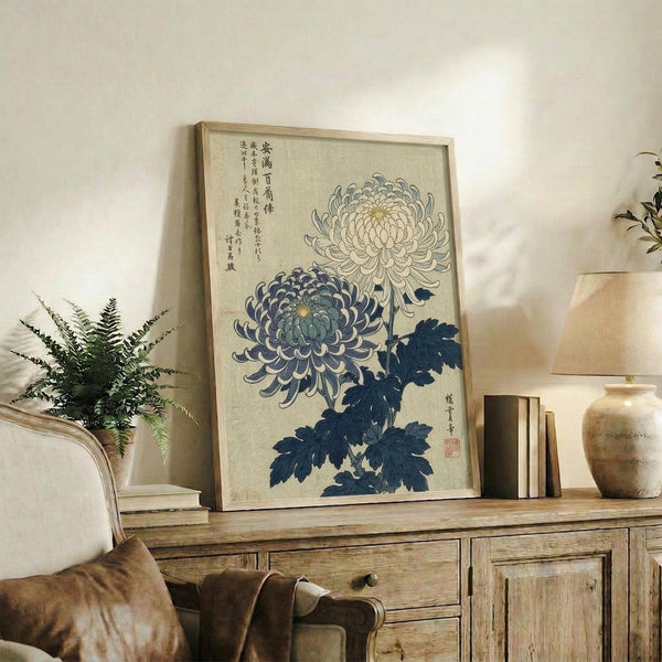 May include: Framed vintage Japanese botanical print featuring blue and white chrysanthemum flowers with traditional calligraphy, displayed on rustic wooden sideboard alongside fern plant, ceramic vases, and books in neutral-toned living room setting with cream upholstered furniture