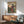 May include: Japanese woodblock print in thin wood frame hanging above mid-century modern credenza with table lamp. Artwork shows woman in colorful kimono with floral patterns standing by wooden gate, cherry blossoms and mountains in background. Room features mint green upholstered chair with wooden arms, round coffee table, white walls with natural sunlight creating geometric shadows.