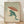May include: Framed artwork showing a green frog holding a rust-colored umbrella in heavy rain, created in Japanese woodblock print style with vertical rain lines filling the background. The frog sits on cream-colored ground with water puddles around its feet. Japanese characters and red seal visible in bottom left corner. Natural wood frame displayed against white wall on light wooden floor.