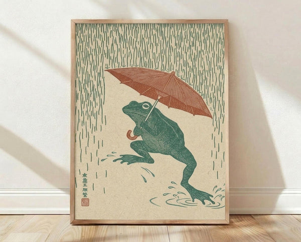 May include: Framed artwork showing a green frog holding a rust-colored umbrella in heavy rain, created in Japanese woodblock print style with vertical rain lines filling the background. The frog sits on cream-colored ground with water puddles around its feet. Japanese characters and red seal visible in bottom left corner. Natural wood frame displayed against white wall on light wooden floor.