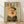 May include: Framed poster print leaning against white wall on light wood floor. Shows orange Pomeranian dog wearing olive green military-style jacket with brass buttons and matching beret on sage green background. Red "DOGUE" text at top mimics fashion magazine cover style. Natural wood frame, portrait orientation, casting soft shadows on textured white wall.