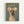 Framed poster showing a small gray dog wearing a cream-colored knit sweater and matching hat with colorful pom-pom ears, photographed against a sage green background. Gold text reading "DOGUE" appears at the top in a fashion magazine style. The wooden frame sits against a white wall.