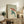 May include: Japanese-style artwork showing a green frog holding a red umbrella in falling rain, displayed in a light wood frame on a rustic wooden sideboard. The print features traditional woodblock print aesthetics with muted colors on cream background. Scene includes fern plant, ceramic vase, books, and neutral-toned furniture in softly lit room setting.