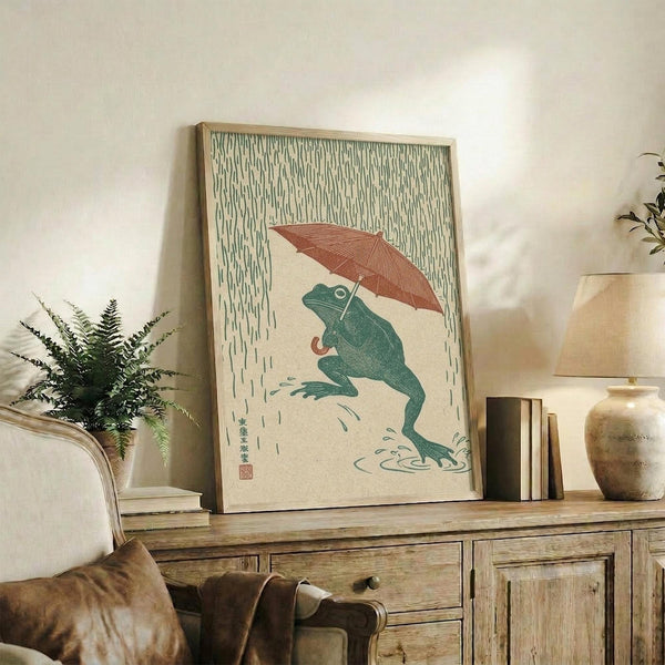 May include: Japanese-style artwork showing a green frog holding a red umbrella in falling rain, displayed in a light wood frame on a rustic wooden sideboard. The print features traditional woodblock print aesthetics with muted colors on cream background. Scene includes fern plant, ceramic vase, books, and neutral-toned furniture in softly lit room setting.