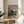 May include: Japanese woodblock print artwork displayed in thin wooden frame on round wooden side table next to beige upholstered chair. Print shows traditional temple building with pink cherry blossoms, small figures in colorful robes walking on path, set against turquoise sky and green landscape. Soft natural lighting creates shadows across white wall. Plant leaves visible in corner, creating serene interior styling scene.