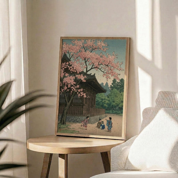 May include: Japanese woodblock print artwork displayed in thin wooden frame on round wooden side table next to beige upholstered chair. Print shows traditional temple building with pink cherry blossoms, small figures in colorful robes walking on path, set against turquoise sky and green landscape. Soft natural lighting creates shadows across white wall. Plant leaves visible in corner, creating serene interior styling scene.
