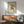 May include: Japanese-style artwork showing a green frog holding a reddish-brown umbrella in falling rain, displayed in a wooden frame above a mid-century modern credenza with white table lamp, next to a teal upholstered armchair with wooden arms in a sunlit room with white walls