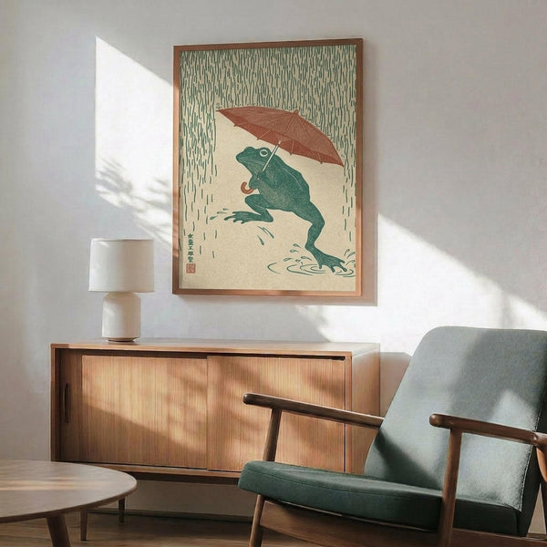 May include: Japanese-style artwork showing a green frog holding a reddish-brown umbrella in falling rain, displayed in a wooden frame above a mid-century modern credenza with white table lamp, next to a teal upholstered armchair with wooden arms in a sunlit room with white walls