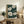 May include: Framed botanical wall art displaying white poppies with detailed petals and brown centers against a dark teal background dotted with small white specks. The wooden-framed artwork leans on a rustic credenza alongside books, ceramic vases, and a fern plant. A beige armchair with leather throw is partially visible in the cozy, neutral-toned room setting.