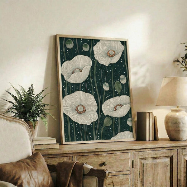 May include: Framed botanical wall art displaying white poppies with detailed petals and brown centers against a dark teal background dotted with small white specks. The wooden-framed artwork leans on a rustic credenza alongside books, ceramic vases, and a fern plant. A beige armchair with leather throw is partially visible in the cozy, neutral-toned room setting.