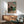 May include: Japanese woodblock print artwork displayed in modern living room with mid-century furniture. The framed poster shows a traditional temple building with pink cherry blossoms and small figures in colorful robes beneath the tree. Artwork hangs above a wood credenza with table lamp, next to a sage green upholstered armchair with wooden arms. Soft natural lighting creates geometric shadows on white walls.