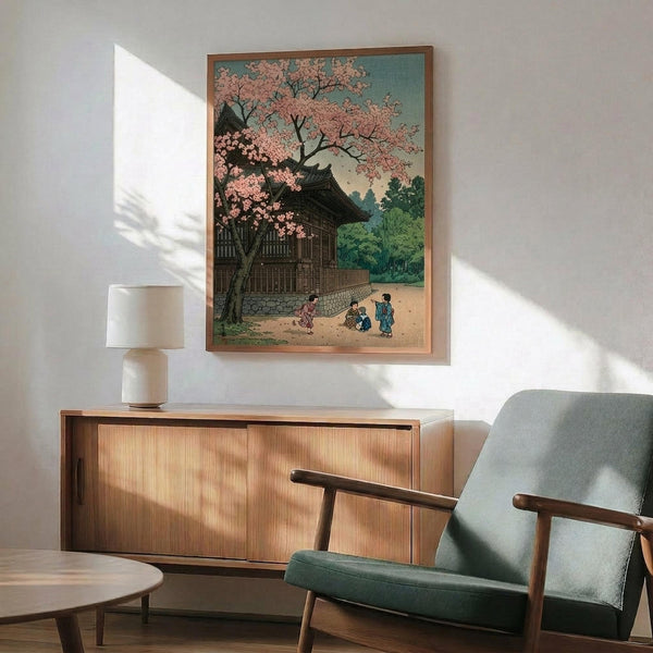 May include: Japanese woodblock print artwork displayed in modern living room with mid-century furniture. The framed poster shows a traditional temple building with pink cherry blossoms and small figures in colorful robes beneath the tree. Artwork hangs above a wood credenza with table lamp, next to a sage green upholstered armchair with wooden arms. Soft natural lighting creates geometric shadows on white walls.