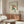May include: Japanese-style artwork featuring a green frog holding a reddish-brown umbrella in falling rain, displayed in a wooden frame above a mid-century modern walnut credenza. The print shows traditional woodblock print styling with vertical rain lines and Japanese characters. Room setting includes a cream upholstered chair, glass-top coffee table, and white table lamp on neutral walls.