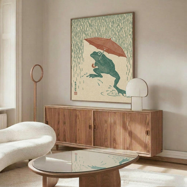 May include: Japanese-style artwork featuring a green frog holding a reddish-brown umbrella in falling rain, displayed in a wooden frame above a mid-century modern walnut credenza. The print shows traditional woodblock print styling with vertical rain lines and Japanese characters. Room setting includes a cream upholstered chair, glass-top coffee table, and white table lamp on neutral walls.