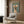 May include: A framed poster featuring an Italian Greyhound wearing a cream knit sweater with colorful pom-pom ear warmers on a blue-green background with "DOGUE" text in yellow letters. The artwork is displayed above a beige upholstered armchair with geometric pillow in a modern living room with wood paneled walls, floor lamp, and side table.