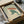 May include: Japanese-style art print featuring a green frog holding a reddish-brown umbrella in falling rain, displayed in a black frame inside an open cardboard shipping box with protective tissue paper and wooden dowels, photographed on a white surface with partial view of a dark glass object in the background