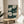 May include: Framed botanical art print displaying white poppies with detailed centers against deep teal background with small white dots, displayed on round wooden side table next to beige upholstered chair in sunlit room with plant shadows on cream wall