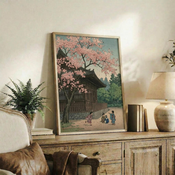May include: Framed Japanese woodblock print featuring cherry blossoms over traditional temple buildings with small figures below, displayed on rustic wooden sideboard alongside potted fern, ceramic vase, and books in neutral-toned living room setting