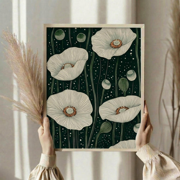 May include: Person holding framed botanical print featuring large white poppies with detailed line work and orange-brown centers against dark green background with white dots. Natural wood frame, cream-colored sleeves visible, dried pampas grass on left side, neutral curtains in soft window lighting.