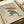 May include: Japanese woodblock-style print displayed in open book or portfolio, showing green frog holding red umbrella in heavy rain. Vintage illustration features detailed line work with teal and rust colors on cream background. Asian characters and red seal stamp visible in corner. Cream ribbon and white papers frame the artistic print.
