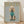 May include: Framed poster print leaning against white wall on wooden floor. Features cream-colored poodle wearing teal turtleneck sweater and matching booties against sage green background. Gold "DOGUE" text at top mimics fashion magazine style. Natural wood frame, soft window lighting creates shadows on textured wall.