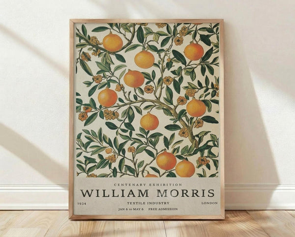 May include: Framed poster with botanical illustration of orange fruit trees and green foliage on cream background, displayed against white textured wall on light wood floor. Vintage-style exhibition poster features William Morris textile industry design from 1934 London centenary show, with elegant typography below ornate fruit and flower pattern in warm oranges and forest greens.